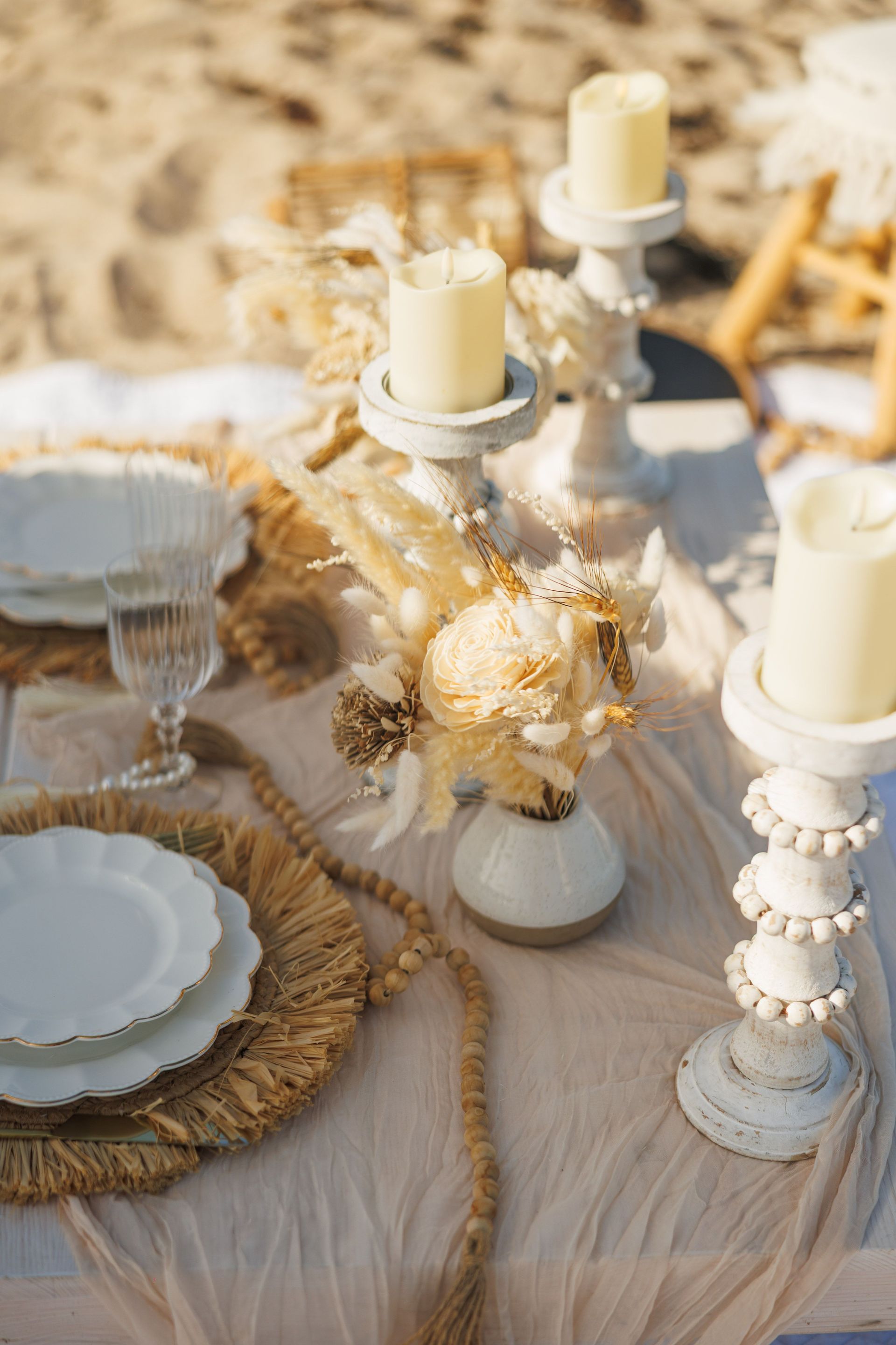 Beach table setting with candles, white plates, and dried flowers; neutral tones.