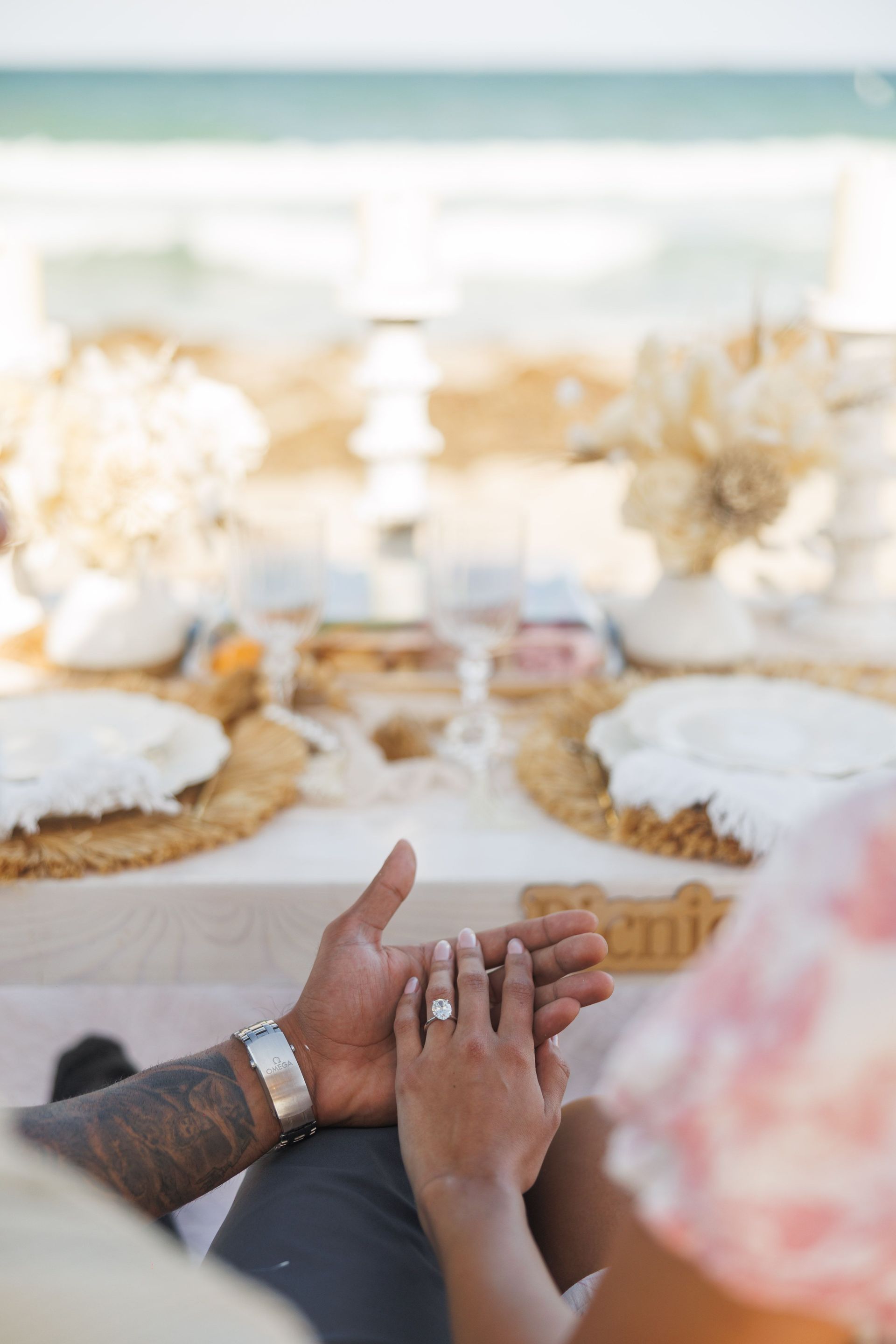 Hands clasped, showing engagement ring, beach setting, table set with white and tan decorations.