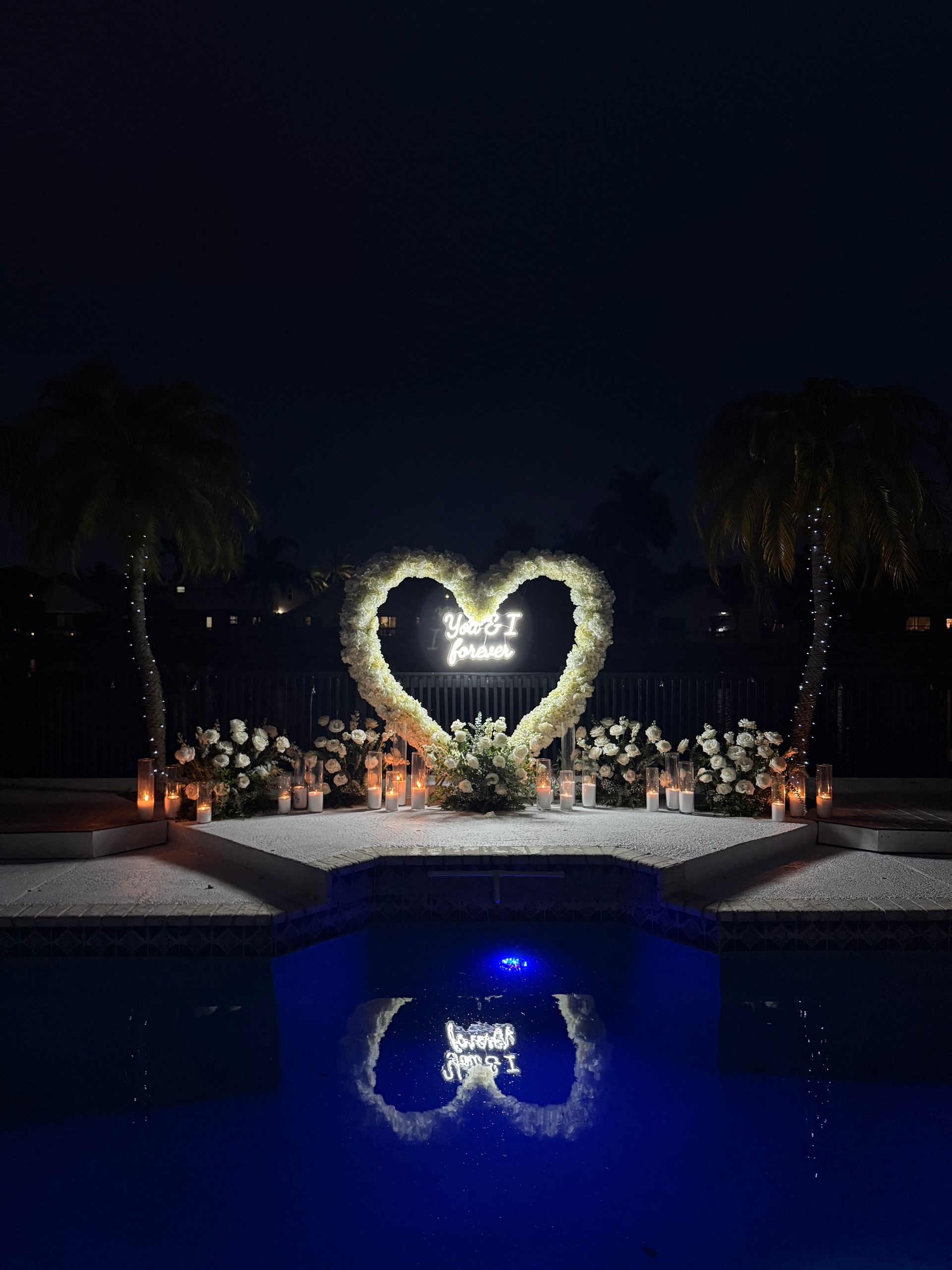 Heart-shaped floral arrangement with glowing sign reflected in a pool at night.