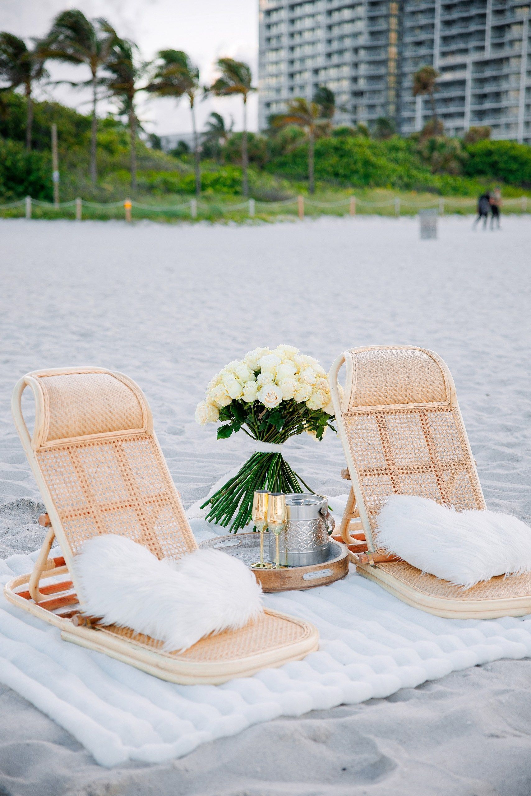 Beach scene with two wicker chairs, flowers, and a candle on a white blanket.