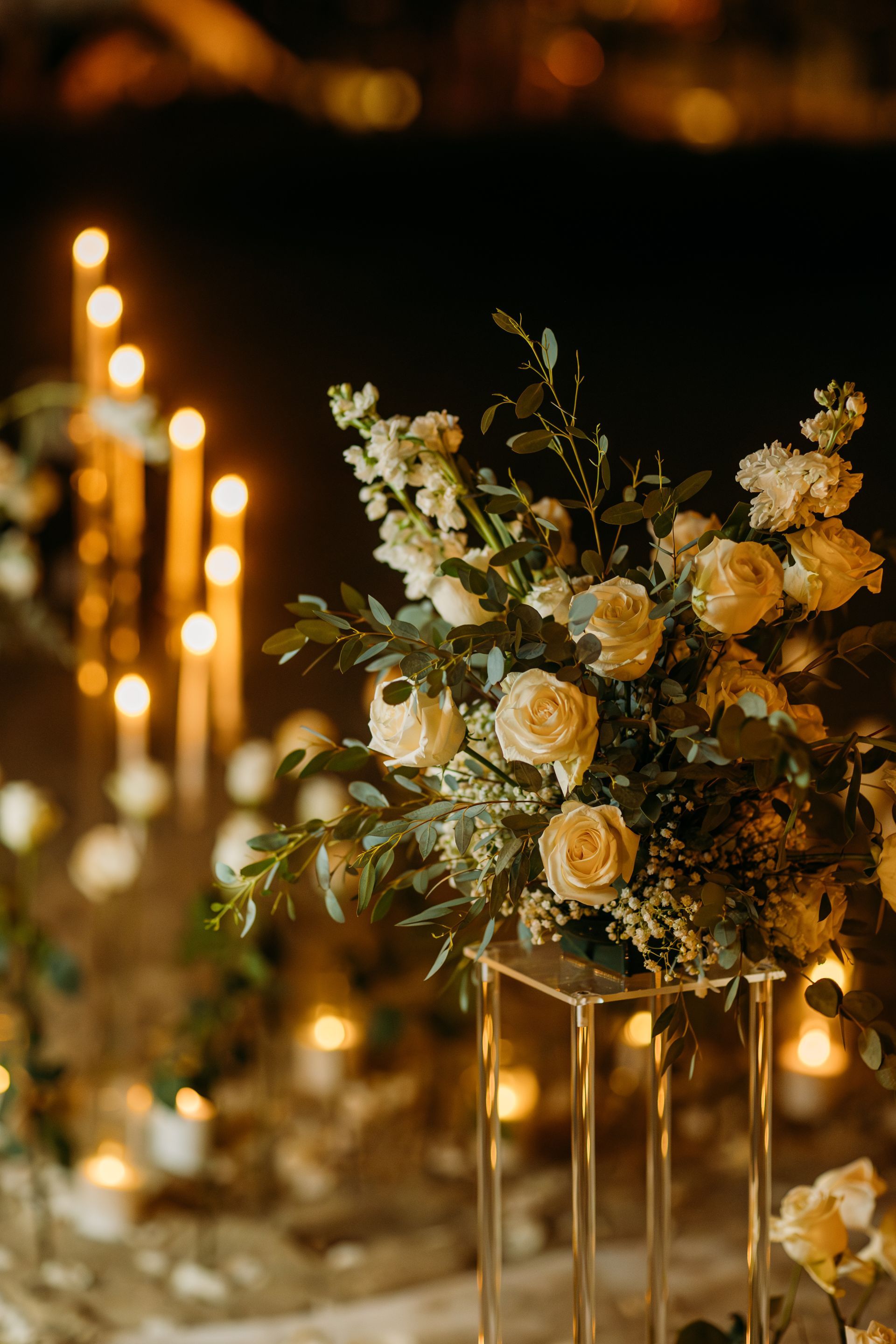 White floral arrangement on a gold stand, surrounded by glowing candles, creating a warm, romantic ambiance.