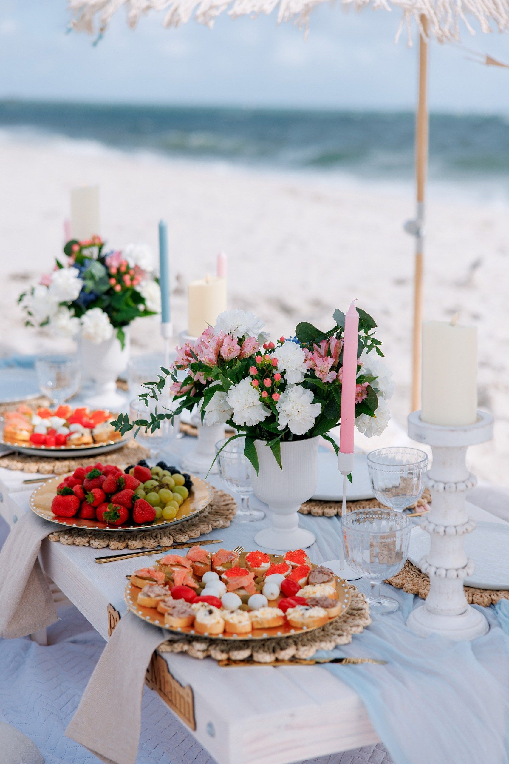 Beach picnic setup with flowers, fruit plates, and candles on a white table, overlooking the ocean.