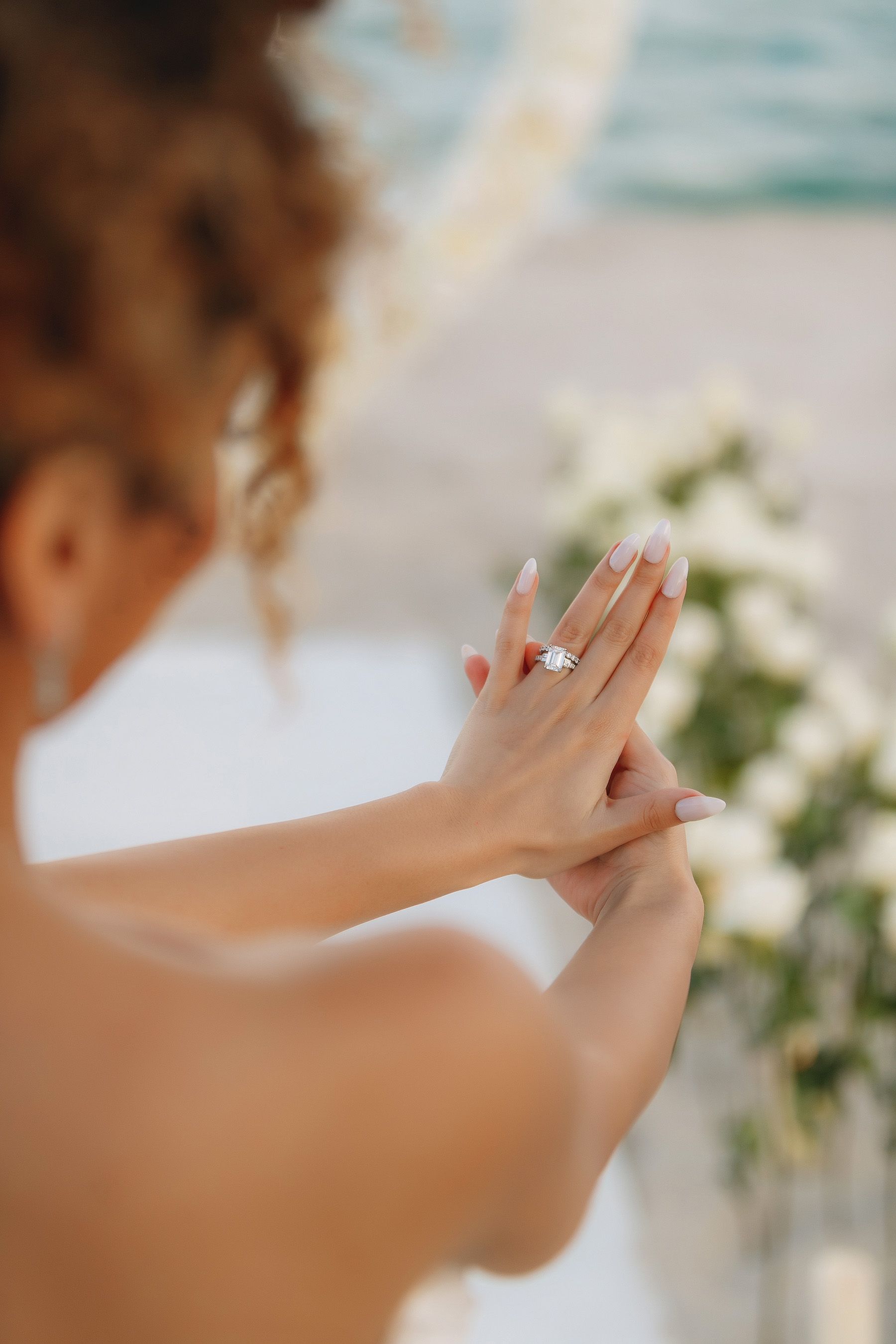Woman admiring engagement ring, white flowers in the background, beach setting.