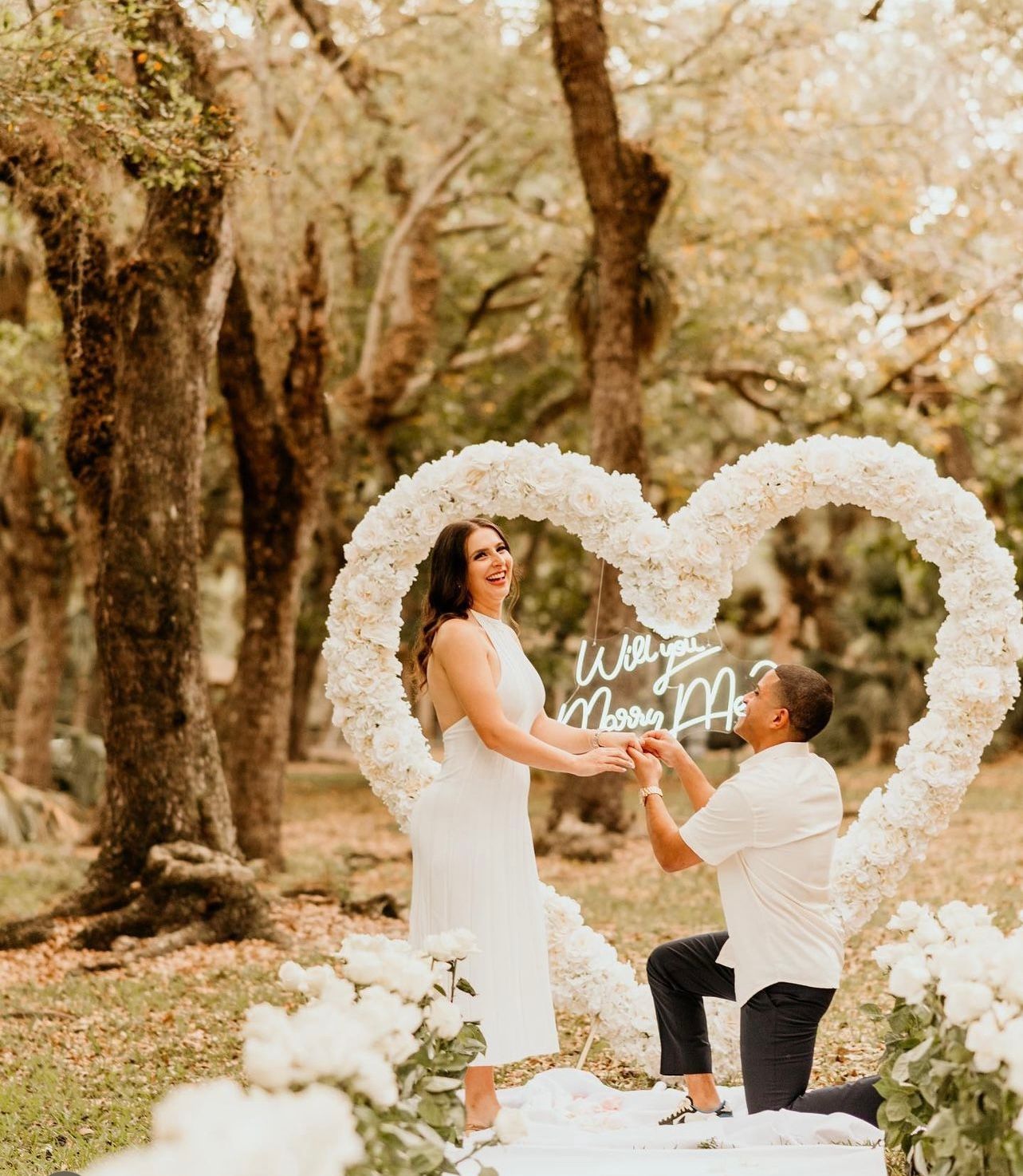 Man proposing to a woman under a floral heart arch. Green trees in background.