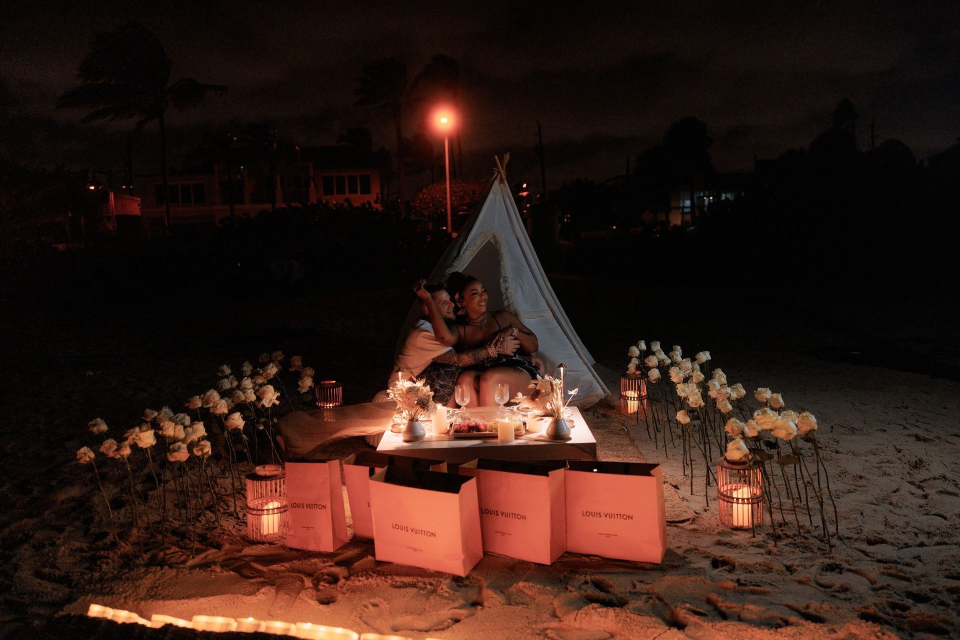 Romantic beach proposal setting with lit candles, flowers, and a couple under a tent at night.