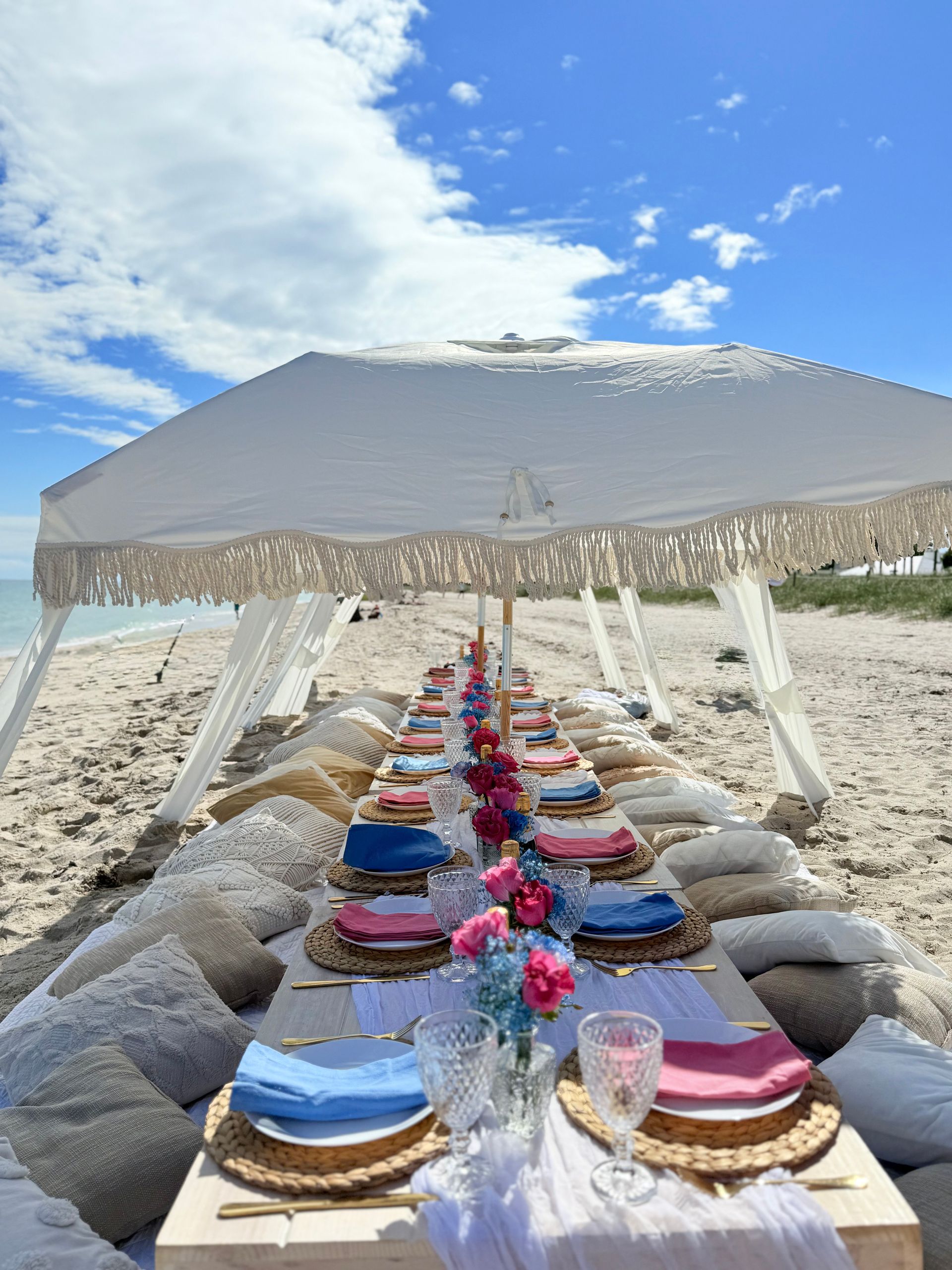 Beach picnic setup with low tables, pillows, and a large umbrella.