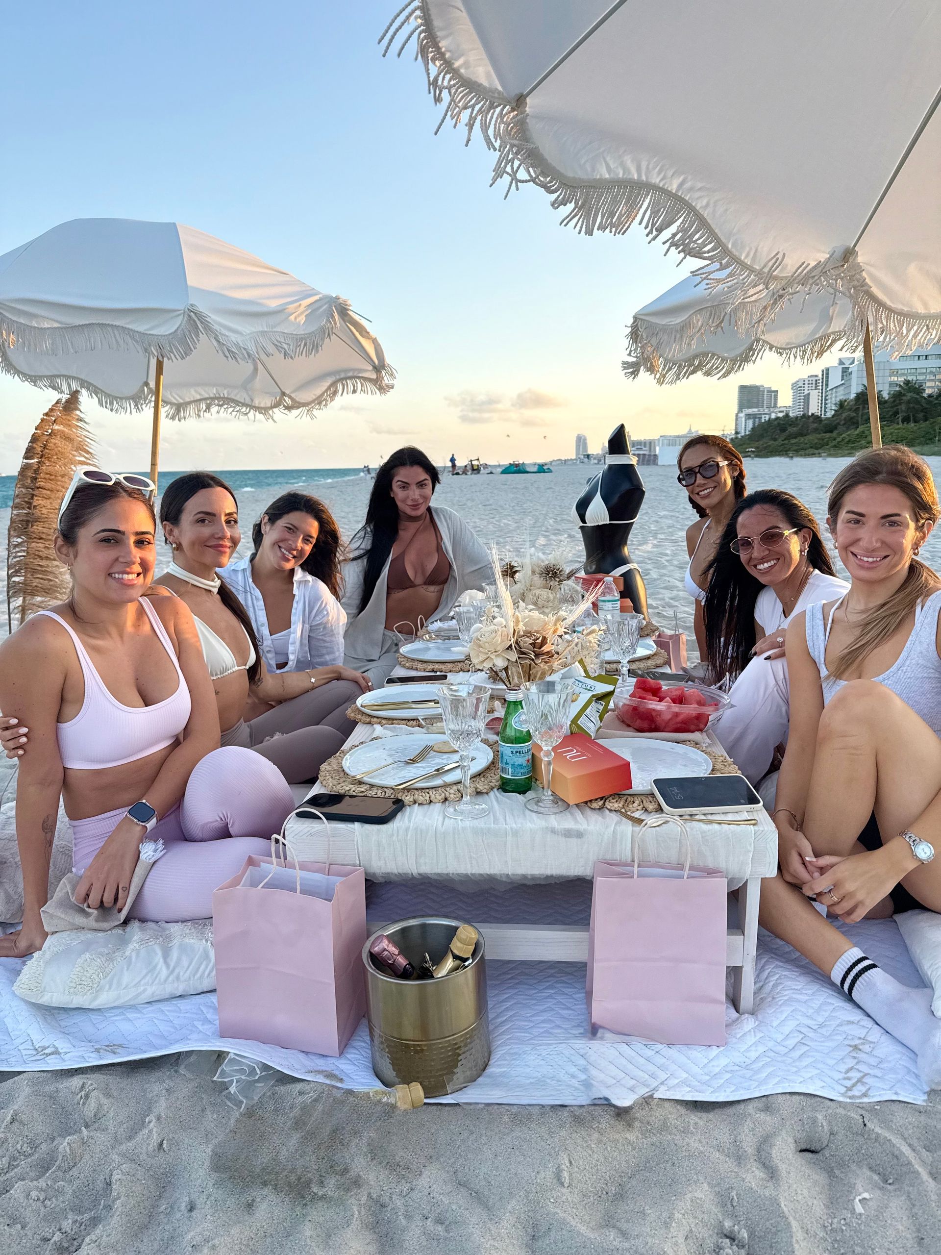 Group of people enjoying a picnic on a beach, under umbrellas. Soft colors, smiling, sunset.
