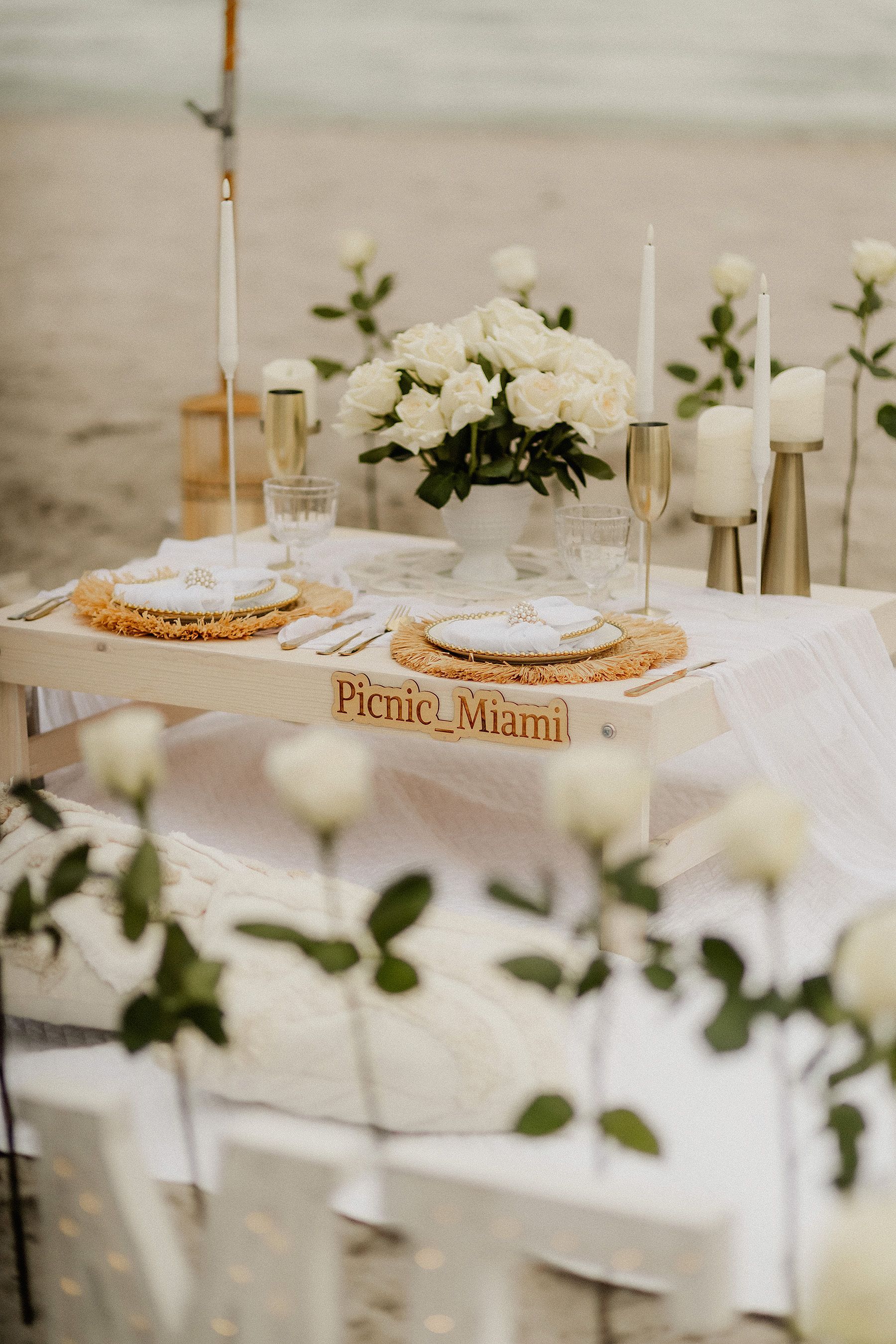 Beach picnic setup: Long wooden table with place settings, pillows, fringed parasol, beach background.