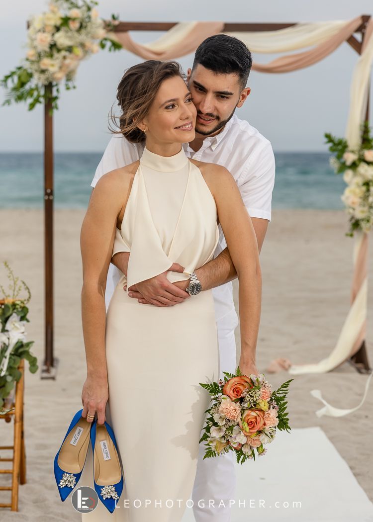 Couple kissing in front of a heart-shaped floral arch on a beach. Wedding setting with ocean backdrop.