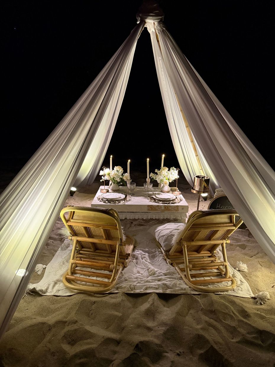 Romantic beach dinner setup with draped fabric canopy, table, flowers, candles, and chairs.