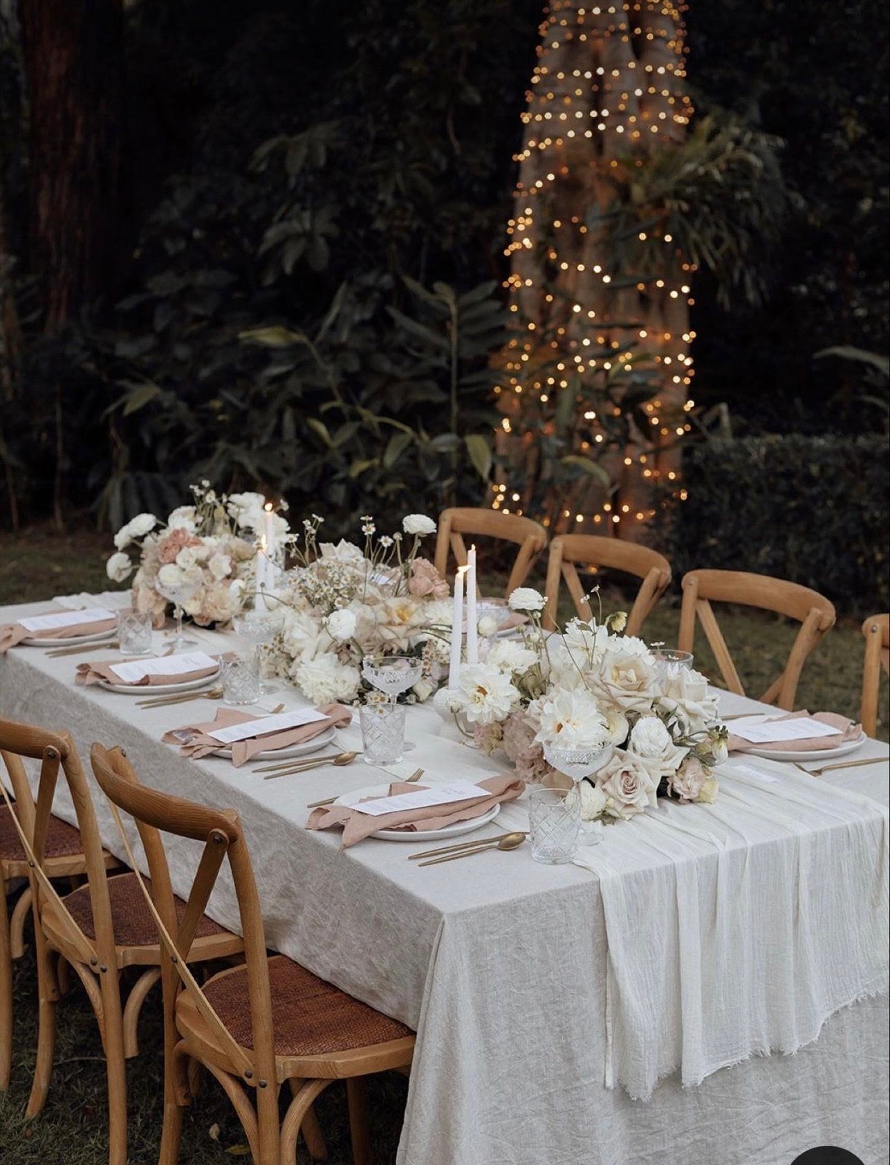 Beach picnic with pink and blue umbrellas, table set with flowers, pillows, and the ocean in the background.