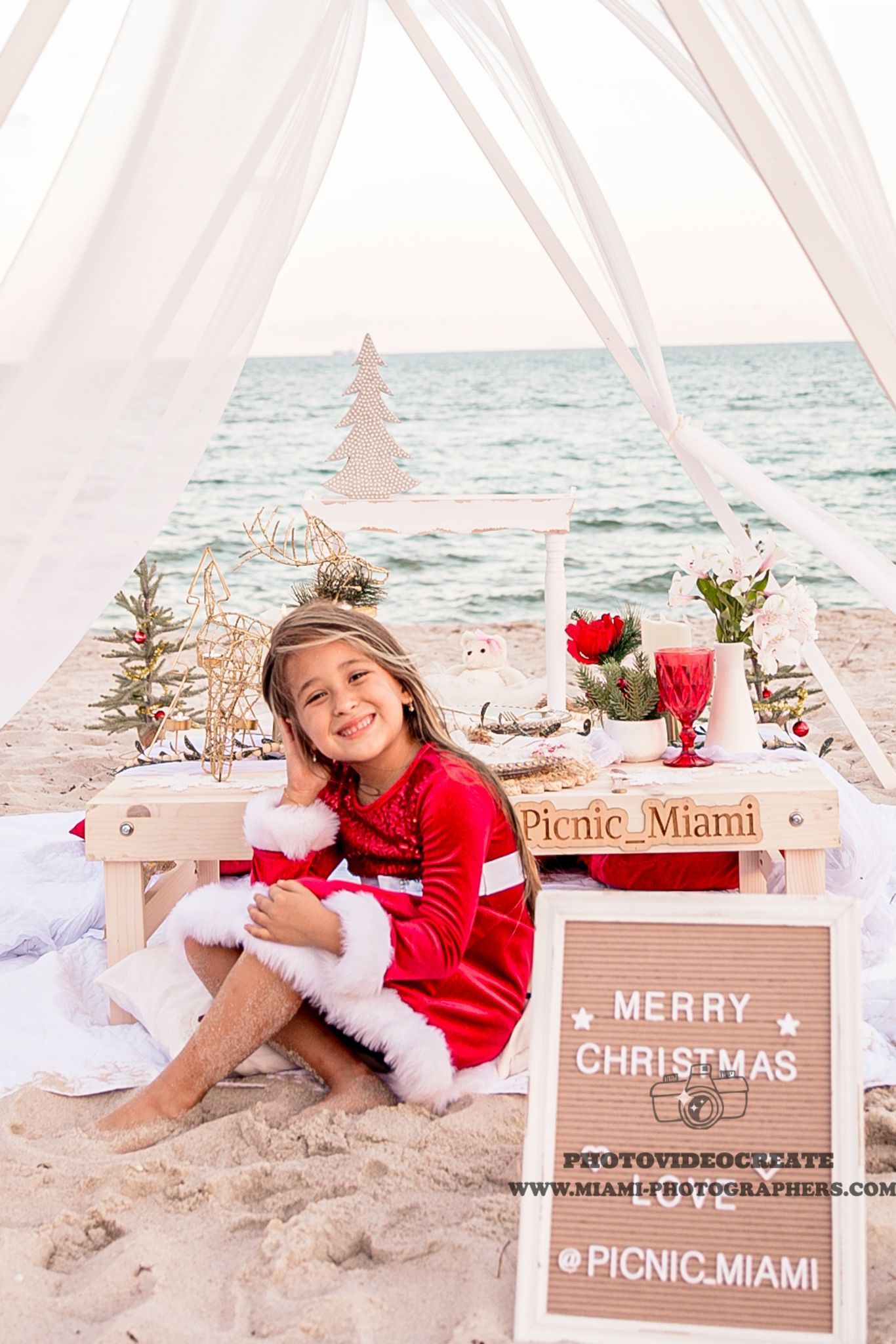Girl in Santa dress sits on a beach, near a picnic table with Christmas decorations and the ocean.