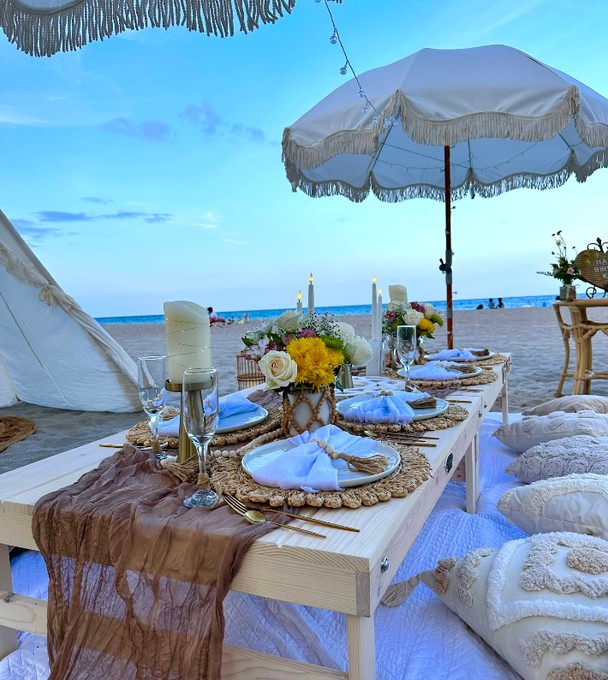 Beach picnic setting with table, decorations, umbrella, pillows, and ocean in the background.