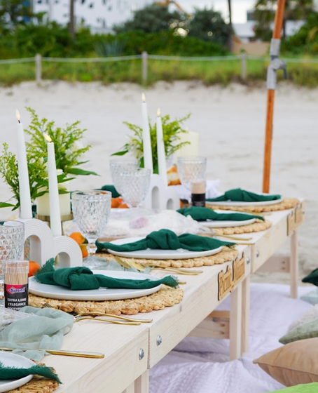 Picnic table set for a beach gathering with candles, green napkins, and woven placemats.