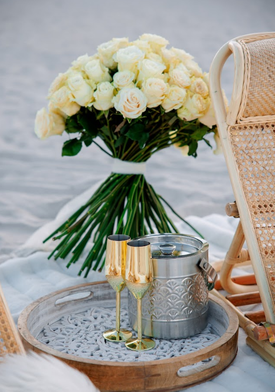 Bouquet of white roses, champagne flutes, and ice bucket on a tray, beach setting.