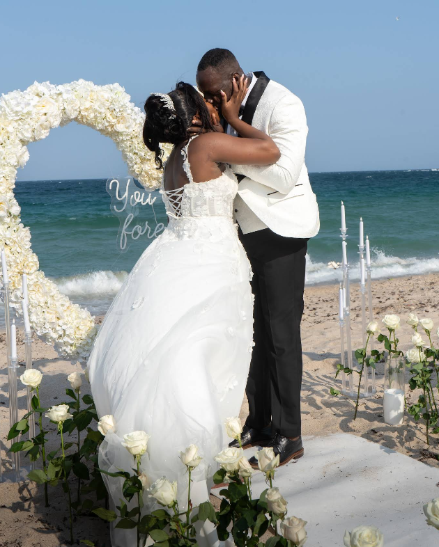 Couple kissing in front of a heart-shaped floral arch on a beach. Wedding setting with ocean backdrop.