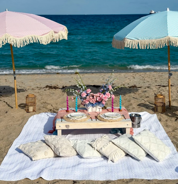 Beach picnic setup with pink and blue umbrellas, food, flowers, and pillows.