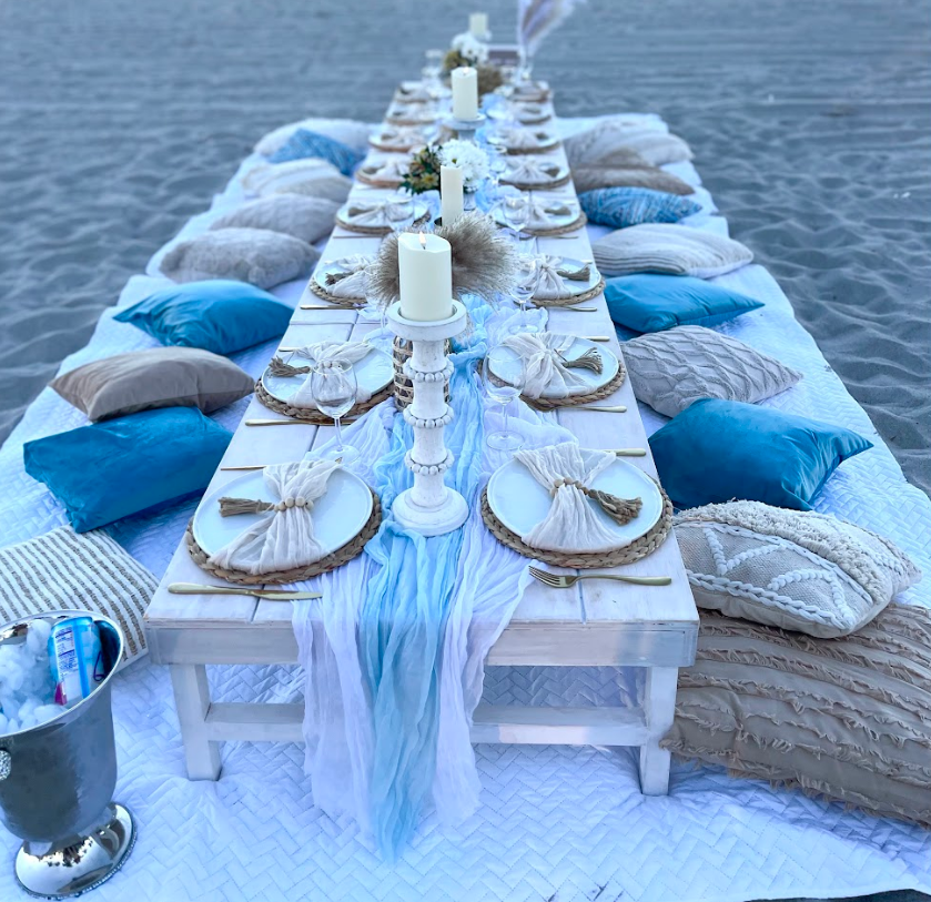 Long table set up on a beach with white, blue decor, candles, pillows, and plates.