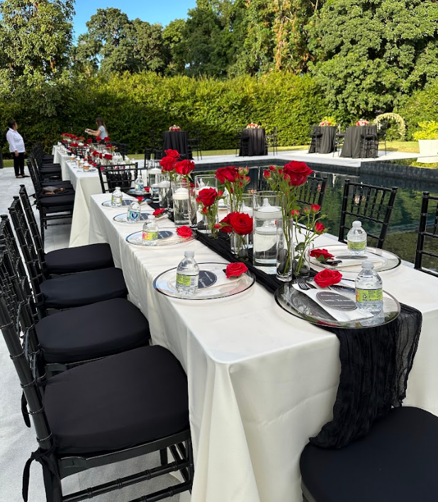 Outdoor dining table set with red roses, black chairs, and a pool in the background.