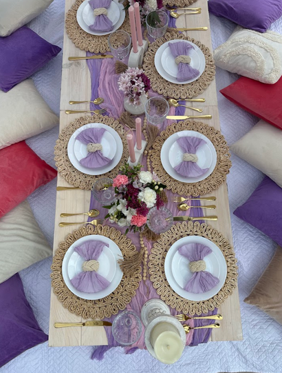Table setting with purple accents, surrounded by pillows. Plates, flowers, candles, and gold cutlery.