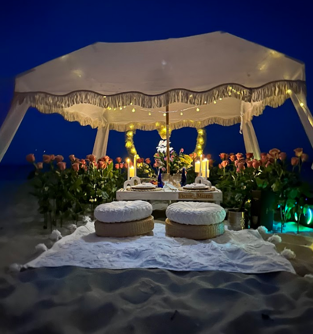 Romantic beach dinner setup with canopy, flowers, candles, and cushions at night.