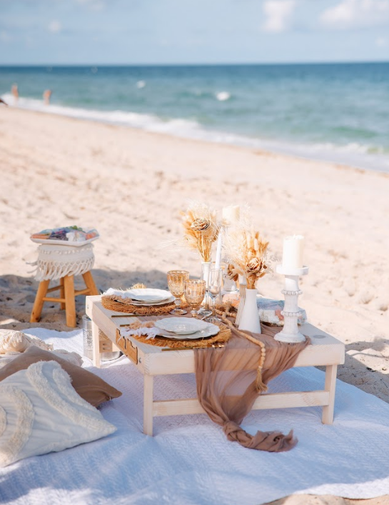 Picnic table set on a sandy beach. Decorated with plates, candles, and a draped fabric, ocean in the background.