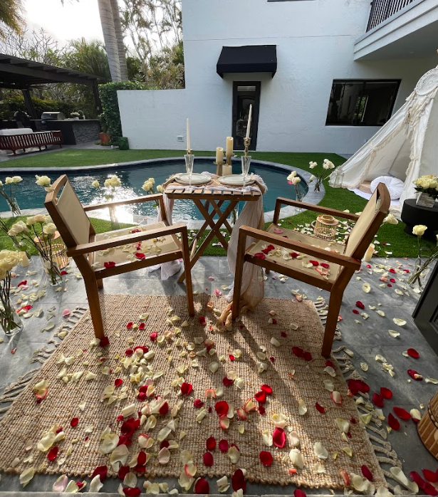 Romantic outdoor table setting with rose petals, chairs, and pool.