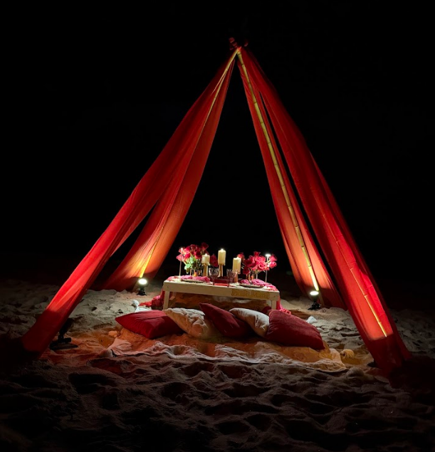 Romantic candlelit picnic on the beach under a draped red canopy.