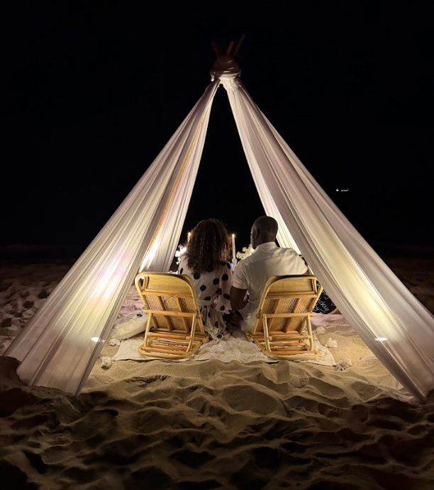 Couple seated under a draped fabric tent on a beach at night; lit by candles, enjoying a romantic dinner.