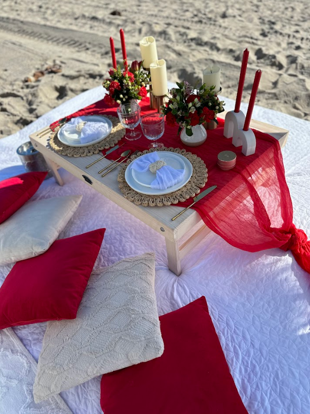 Beach picnic setup with red accents: table, candles, floral arrangements, pillows, on white blanket.