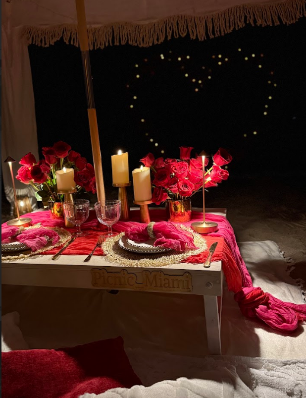 Romantic outdoor dinner setting: red roses, candles, heart-shaped lights, table on the beach.