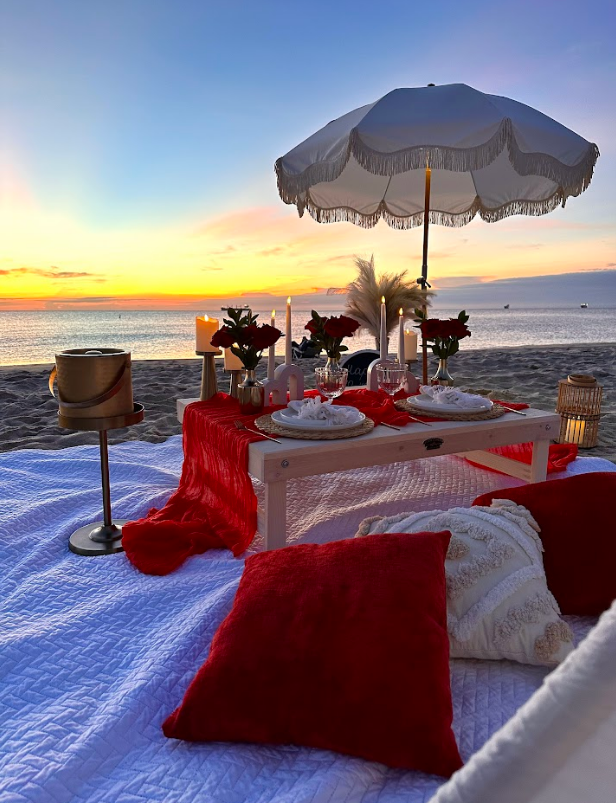 Romantic beach picnic setting at sunset with red and white decor.