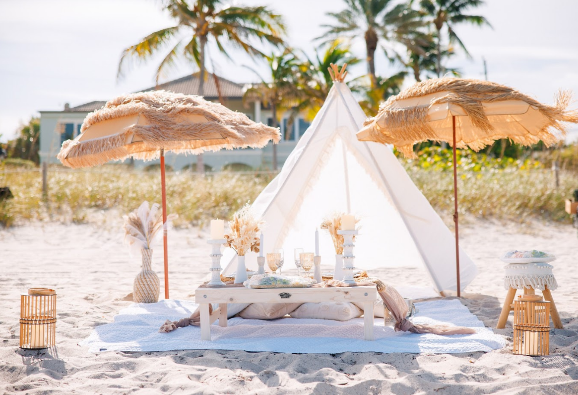Beach picnic setup with parasols, teepee, table, and decorations.