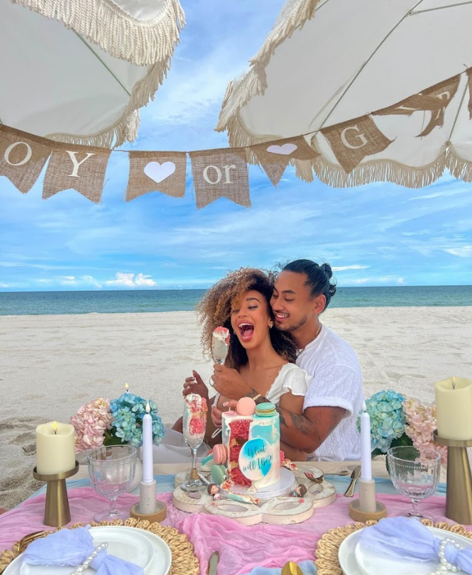 Couple at a beach gender reveal party, celebrating with cake. Pink and blue decorations, banner reads