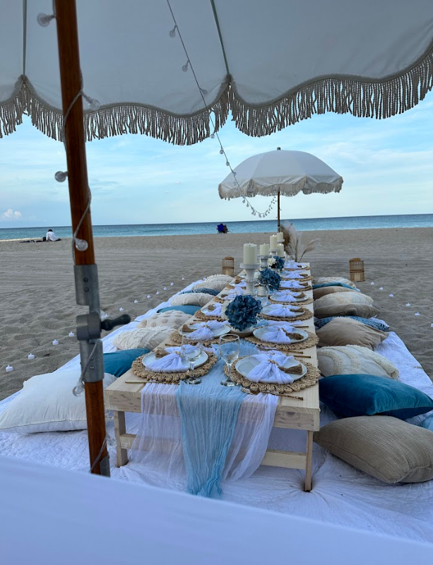 Beach picnic setup with a long table, pillows, and decor in blue and white.