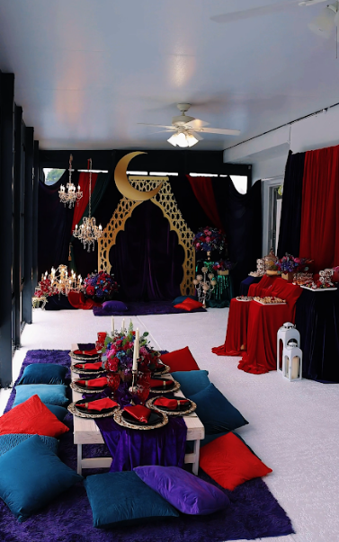 Room with low table set for dining, surrounded by cushions in red, teal, and purple. Backdrop with red/black curtains.