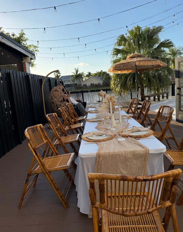 Outdoor dining table set for a gathering. Wooden chairs, white tablecloth, and string lights overhead.