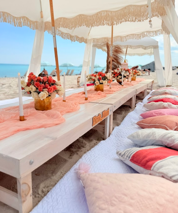 Beach picnic setup with long wooden table, peach accents, flowers, and pillows under a canopy.