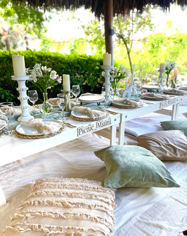 Picnic table set for a meal, with cushions, candles, and flowers, outdoors under a shaded structure.