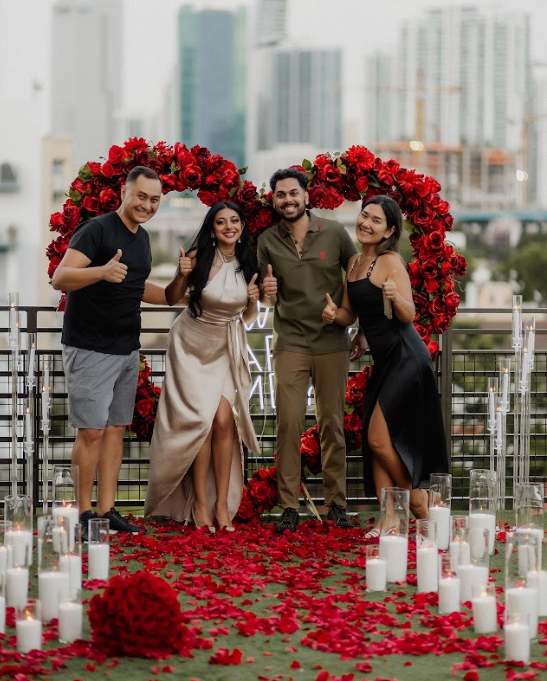 Four people give thumbs-up near a rose-filled heart, likely celebrating an engagement on a rooftop.