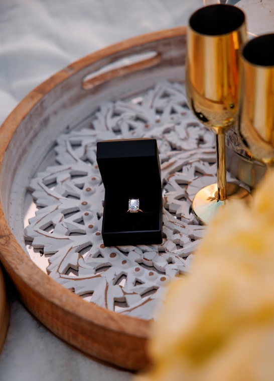 Engagement ring in open black box on decorative tray with gold champagne flutes.