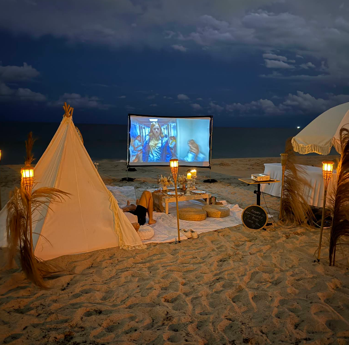 Beach setup with teepee, screen showing video, lit candles, and beach umbrella at dusk.