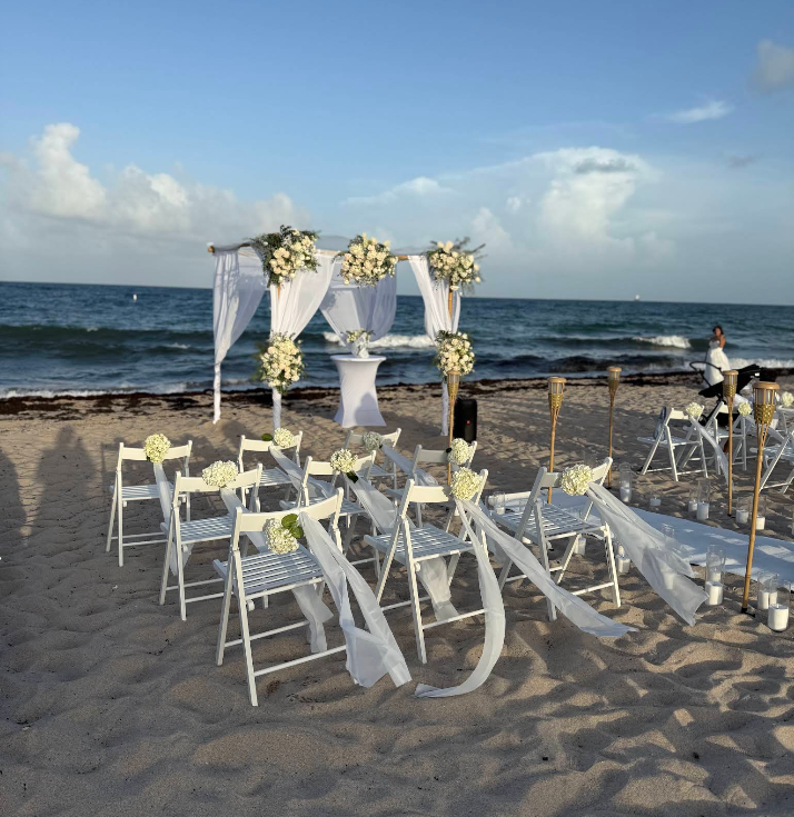Beach wedding setup with white chairs, archway, and decorations against the ocean.