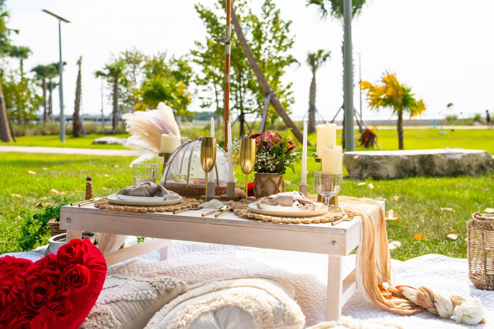 Picnic setup on a white table with candles, flowers, and champagne flutes on a sunny day.