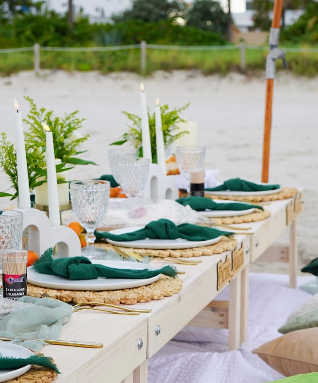 Beach picnic setup: white table, green napkins, candles, place settings, on sand.