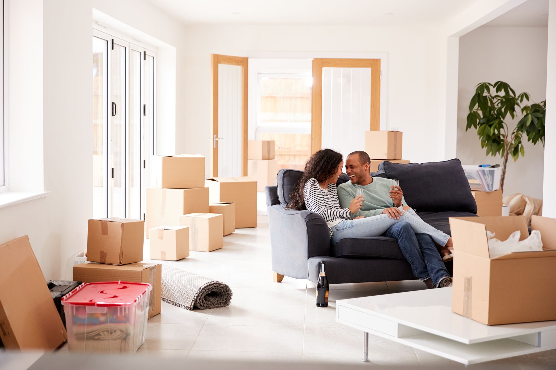 A man and woman are sitting on a couch in a living room surrounded by cardboard boxes.