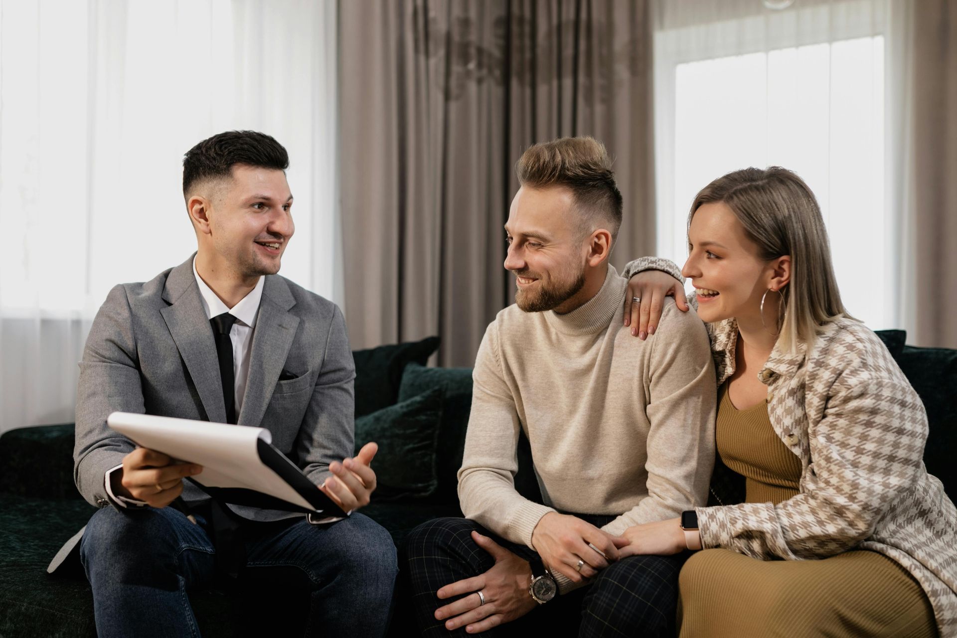 A man in a suit is sitting on a couch talking to a couple. Everyone is smiling.