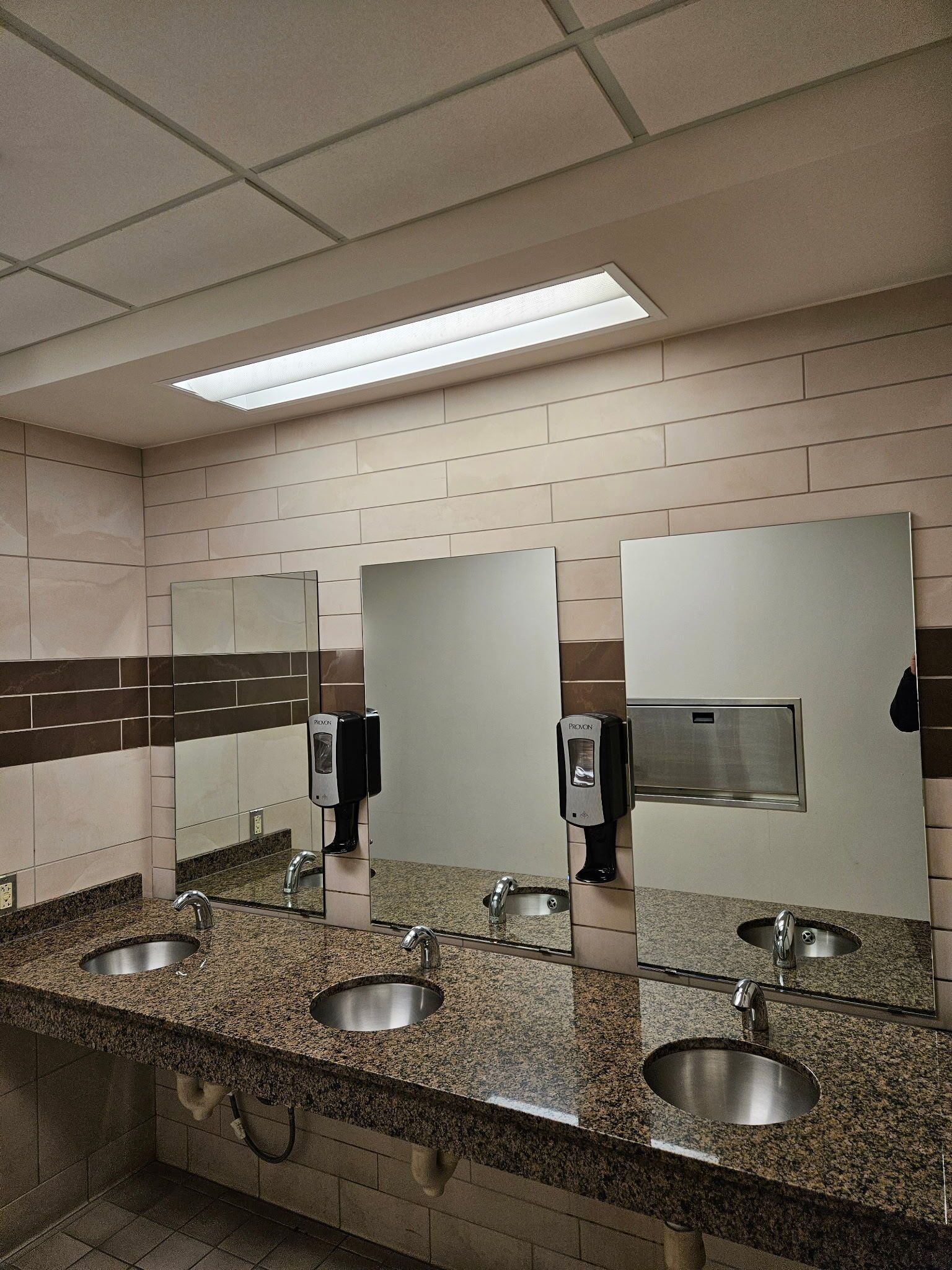 Public restroom with granite counters, three sinks, mirrors, and soap dispensers under fluorescent lights