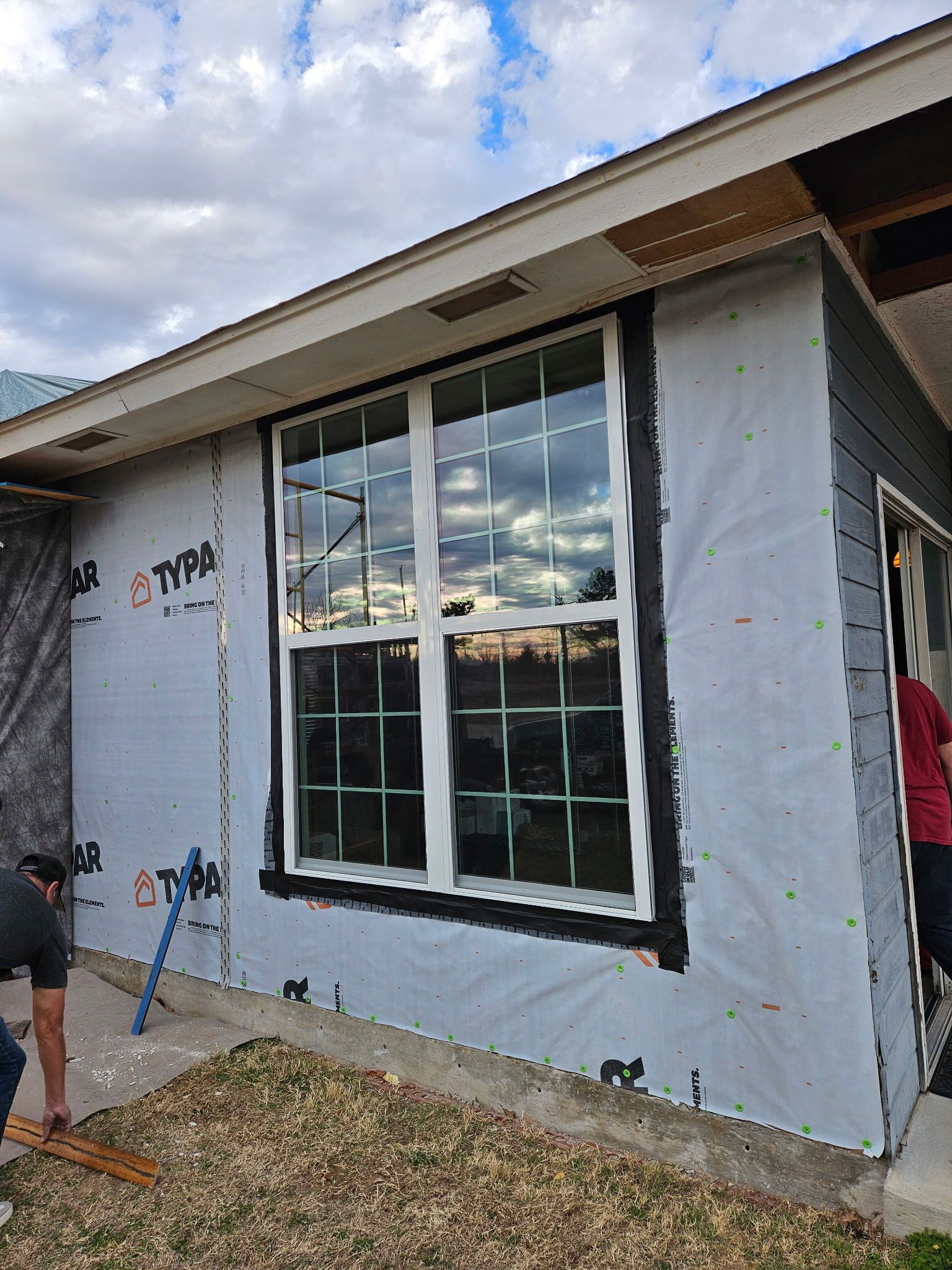 House exterior under renovation with white house wrap, black-trimmed window, and a worker at left