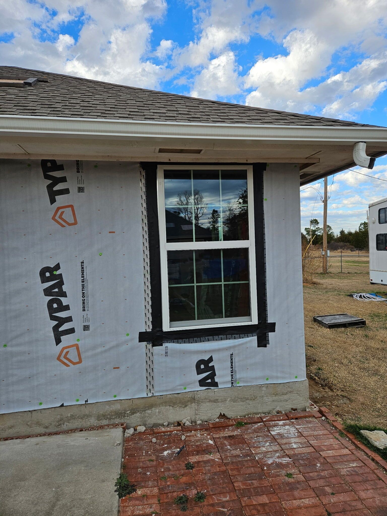 Partially finished house exterior with a window wrapped in black tape and white house wrap under cloudy sky